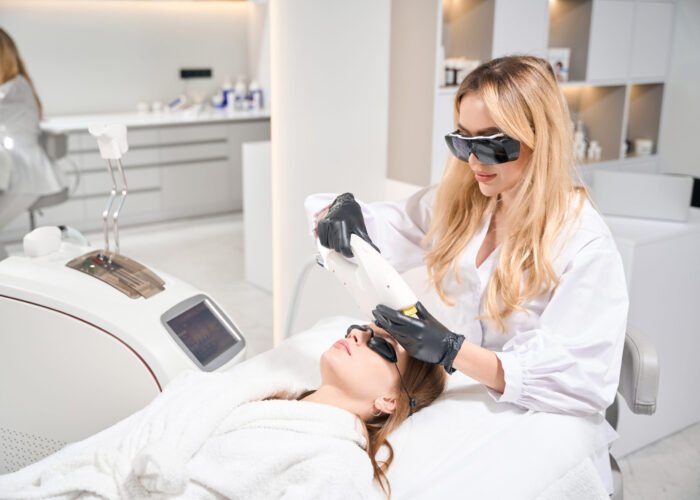 Female beautician doing laser depilation with laser hair removal machine of forehead of young woman in blurred beauty salon. Concept of face skin care
