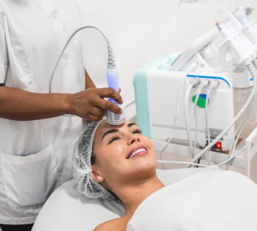 Young woman is lying down smiling and receiving skin treatment from a beautician using a professional device