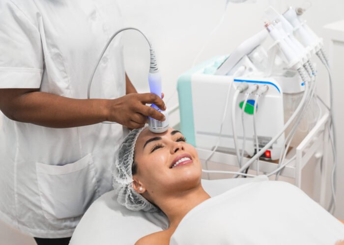 Young woman is lying down smiling and receiving skin treatment from a beautician using a professional device