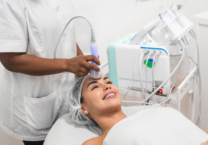 Young woman is lying down smiling and receiving skin treatment from a beautician using a professional device