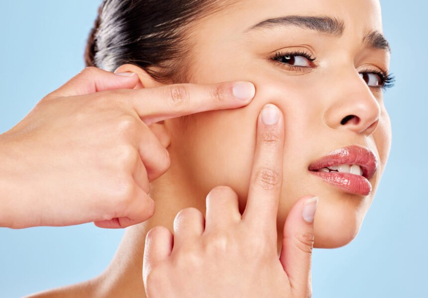 studio-portrait-of-an-attractive-young-woman-squee-2025-04-06-11-50-34-utc Studio portrait of an attractive young woman squeezing a zit on her face against a blue background.